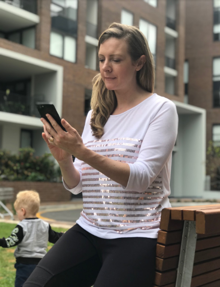 Photo of Kirryn looking at her phone, while her son Bailey runs around in the background.