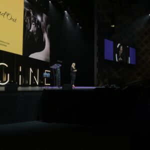 Kirryn is a keynote speaker on a black stage with two screens in the background