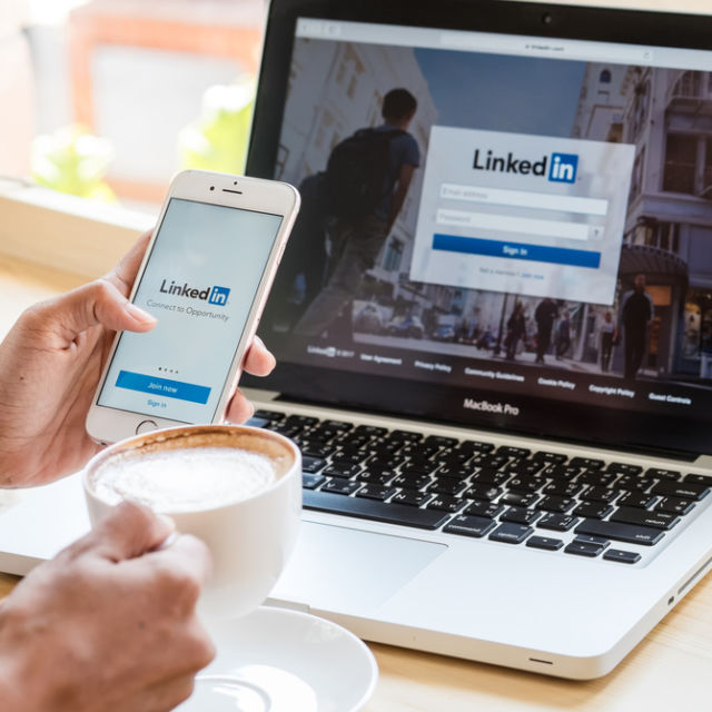 A phone held up in front of a laptop, both with a picture of the Linkedin sign on page